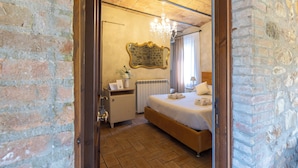 A bedroom with a brick wall, a wooden ceiling, a chandelier, a bed with white bedding, a nightstand, a mirror, and a radiator. - Casale Serena 1912 (Fabro)