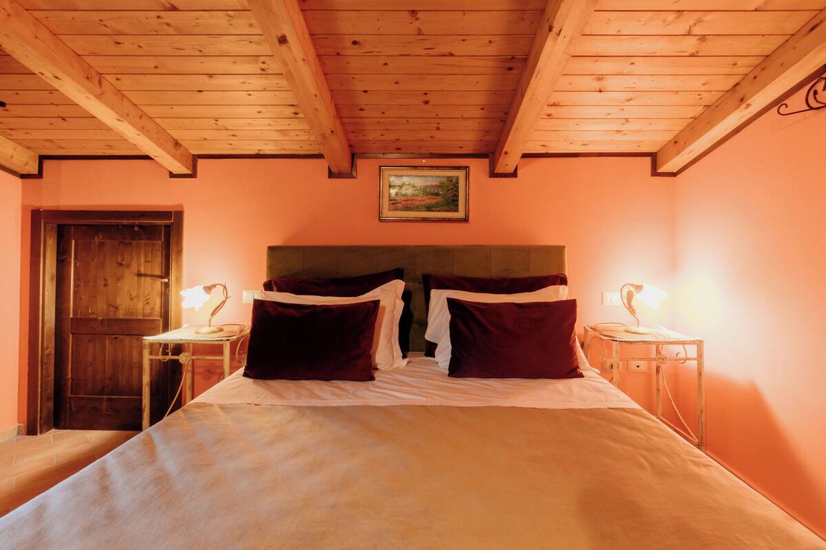 A bedroom with a wooden ceiling, a bed with white and dark red bedding, two bedside tables with lamps, and a painting on the wall.