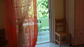 A room with a sliding glass door leading to an outdoor area with a green plant backdrop. A wooden chair and a small table are visible inside the room. - Studio Stars (Zakynthos)