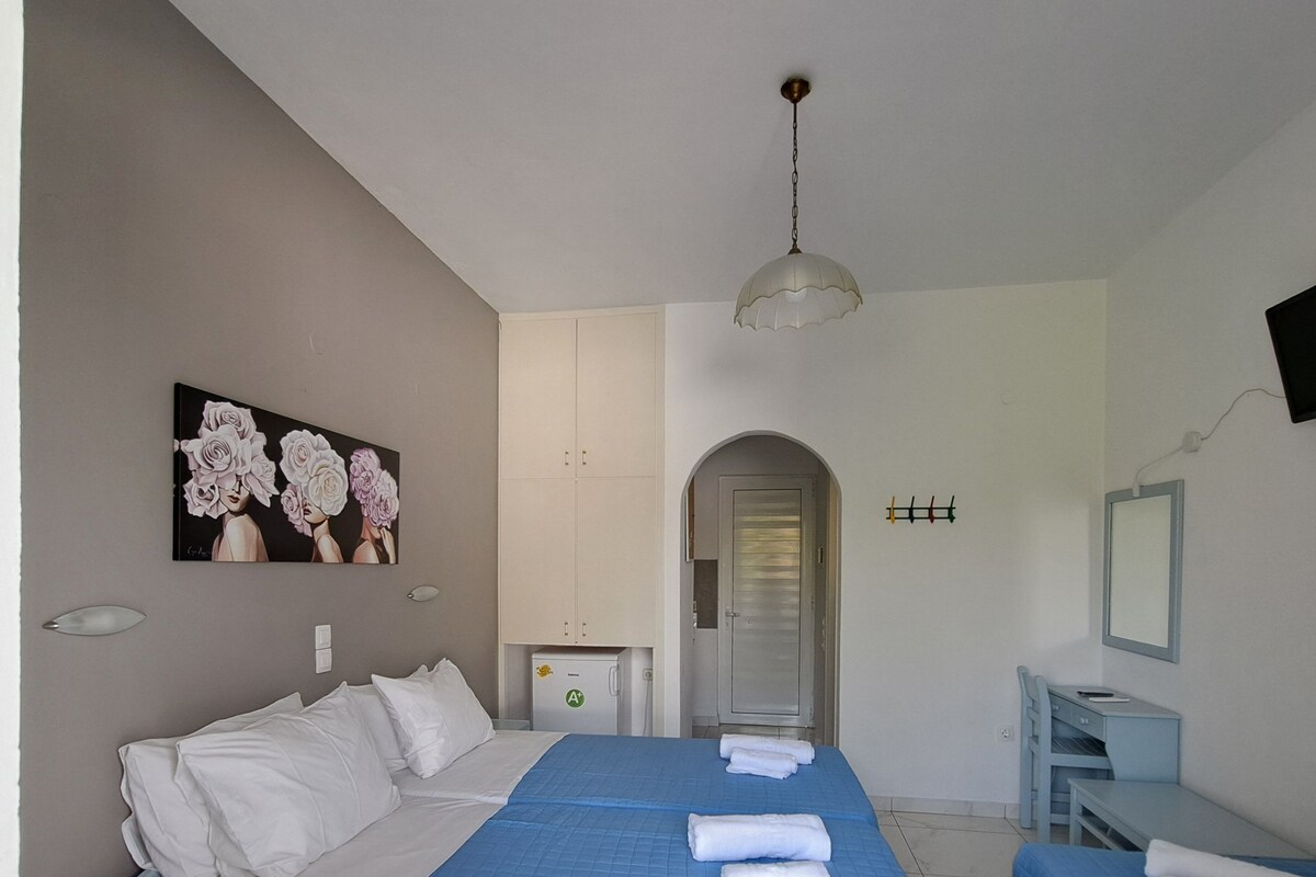 A hotel room with a large bed, a smaller bed, a desk, a chair, a wardrobe, a painting, a ceiling light, and a door leading to a hallway.