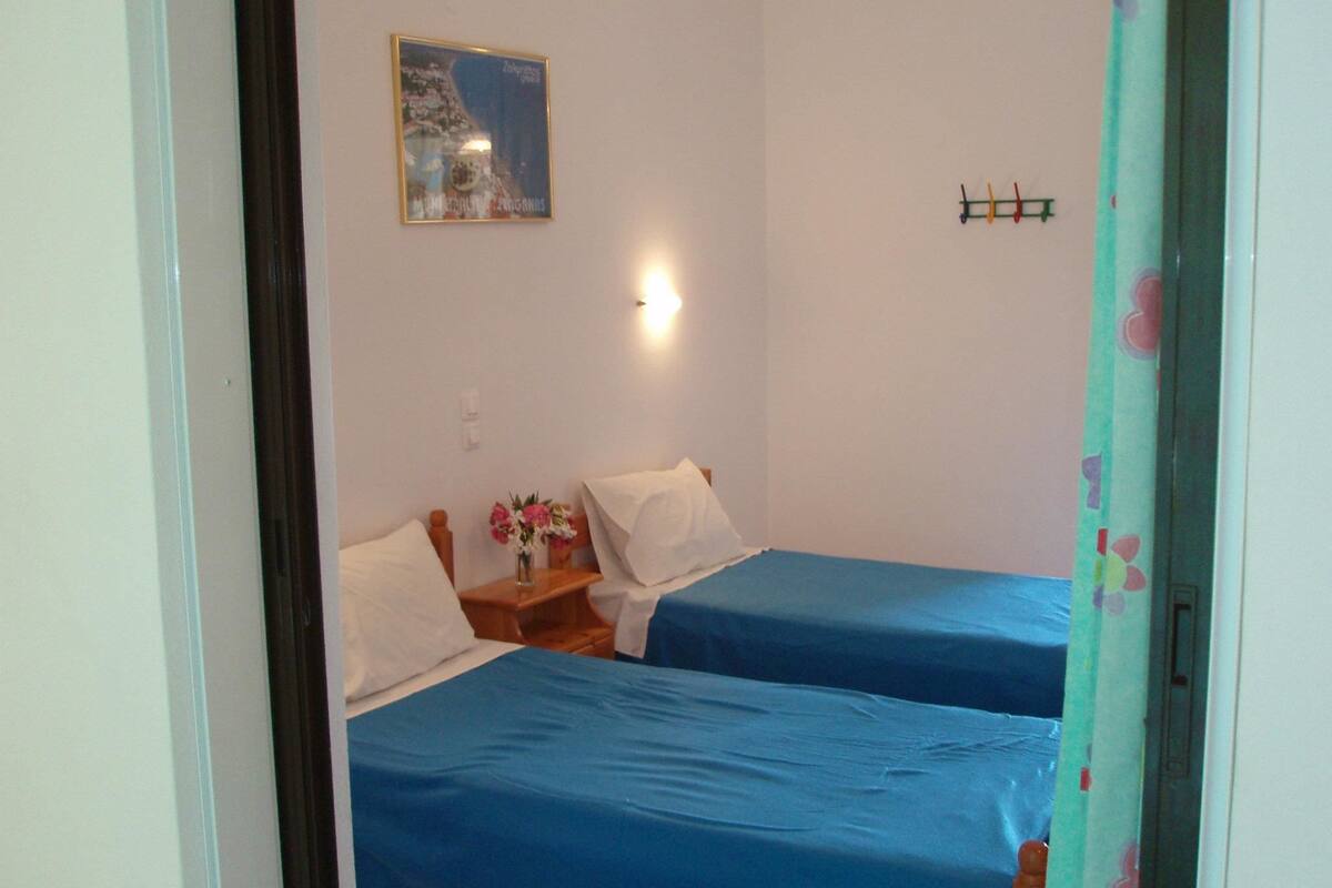 A room with two beds, a nightstand with a vase of flowers, a wall-mounted light, and a framed picture.