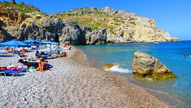 Une plage de galets avec des parasols et des gens qui prennent des bains de soleil, des falaises rocheuses et une eau d’un bleu limpide.