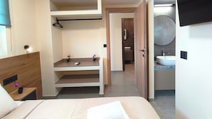 Une chambre moderne avec un lit, une table de chevet sur laquelle se trouve un téléphone, une petite étagère avec deux plantes en pot, et une salle de bain visible à travers une porte ouverte.