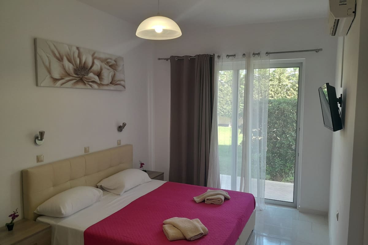Ein Schlafzimmer mit einem Bett, einem Nachttisch, einem Gemälde, einer Deckenleuchte, einem Fenster mit Vorhängen und einem an der Wand montierten Fernseher.