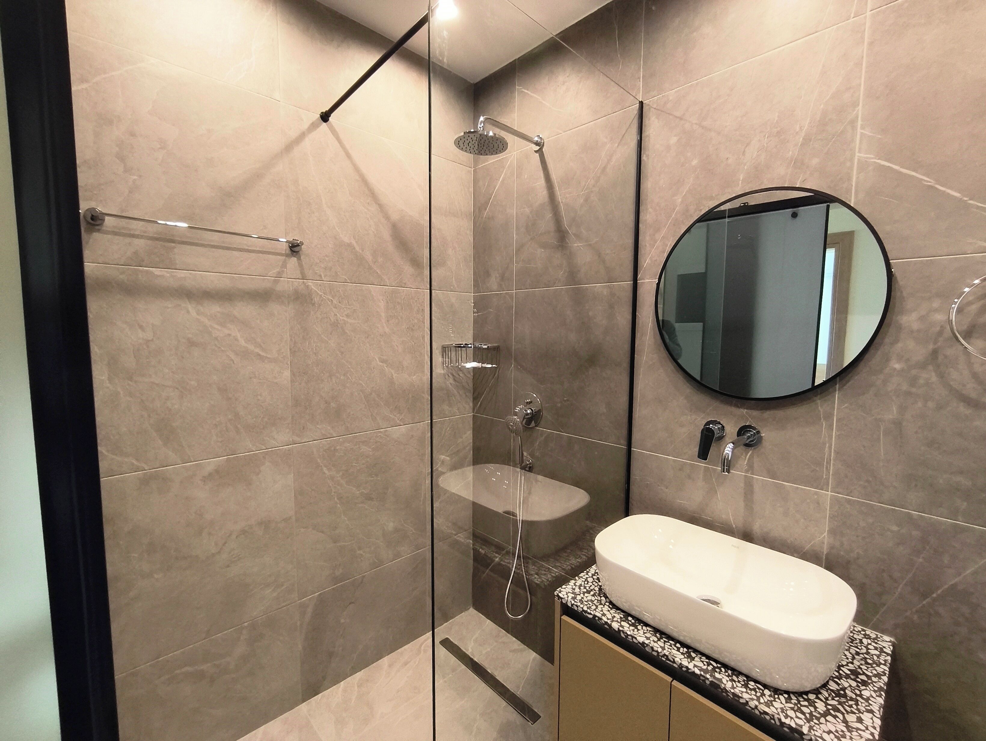 A modern bathroom with a glass-enclosed shower, a round mirror, and a white sink with a mosaic countertop.