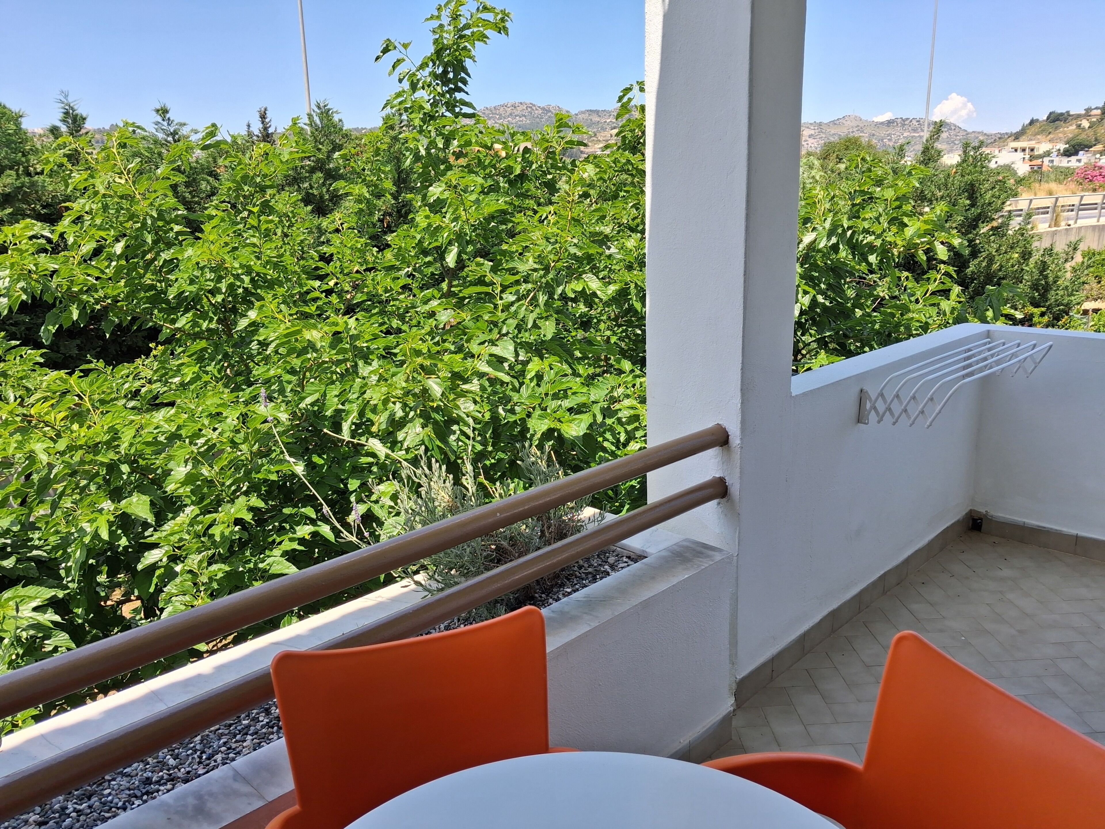 Un balcón con barandilla blanca, sillas naranjas y vistas a la vegetación verde y edificios a lo lejos.