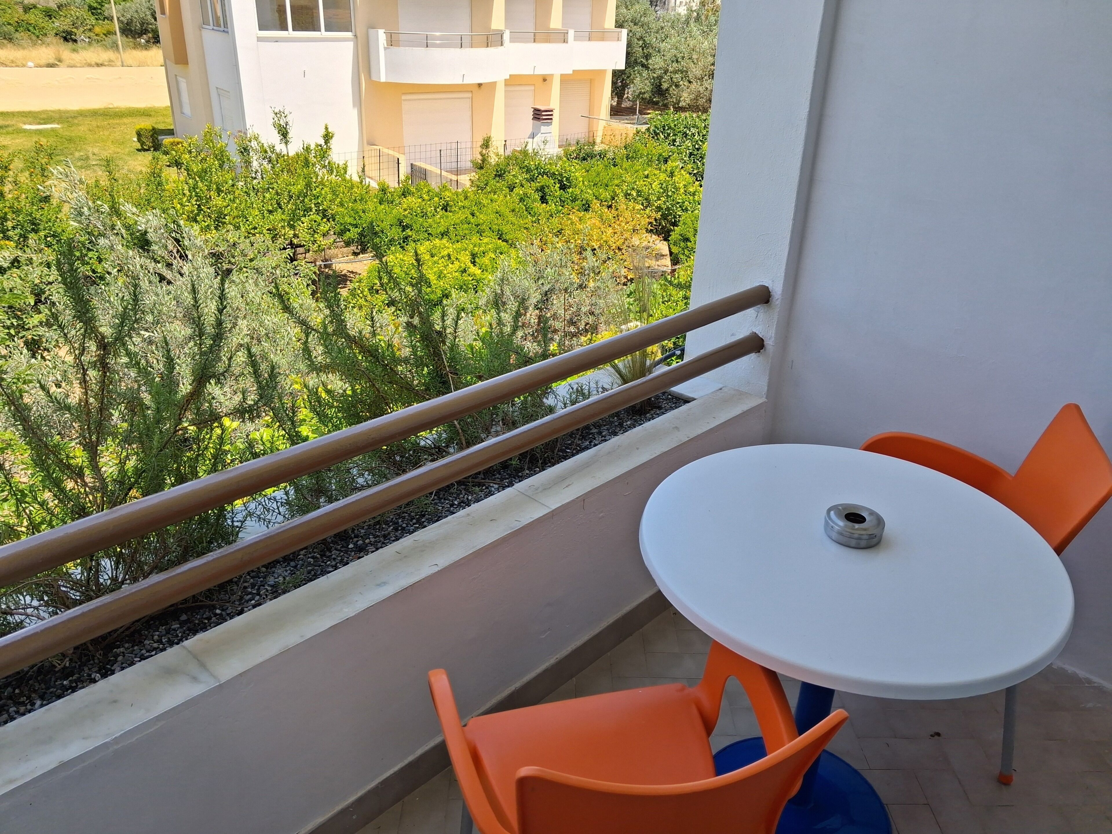 Un balcón con una mesa blanca y dos sillas anaranjadas, con vista a un jardín y edificios.