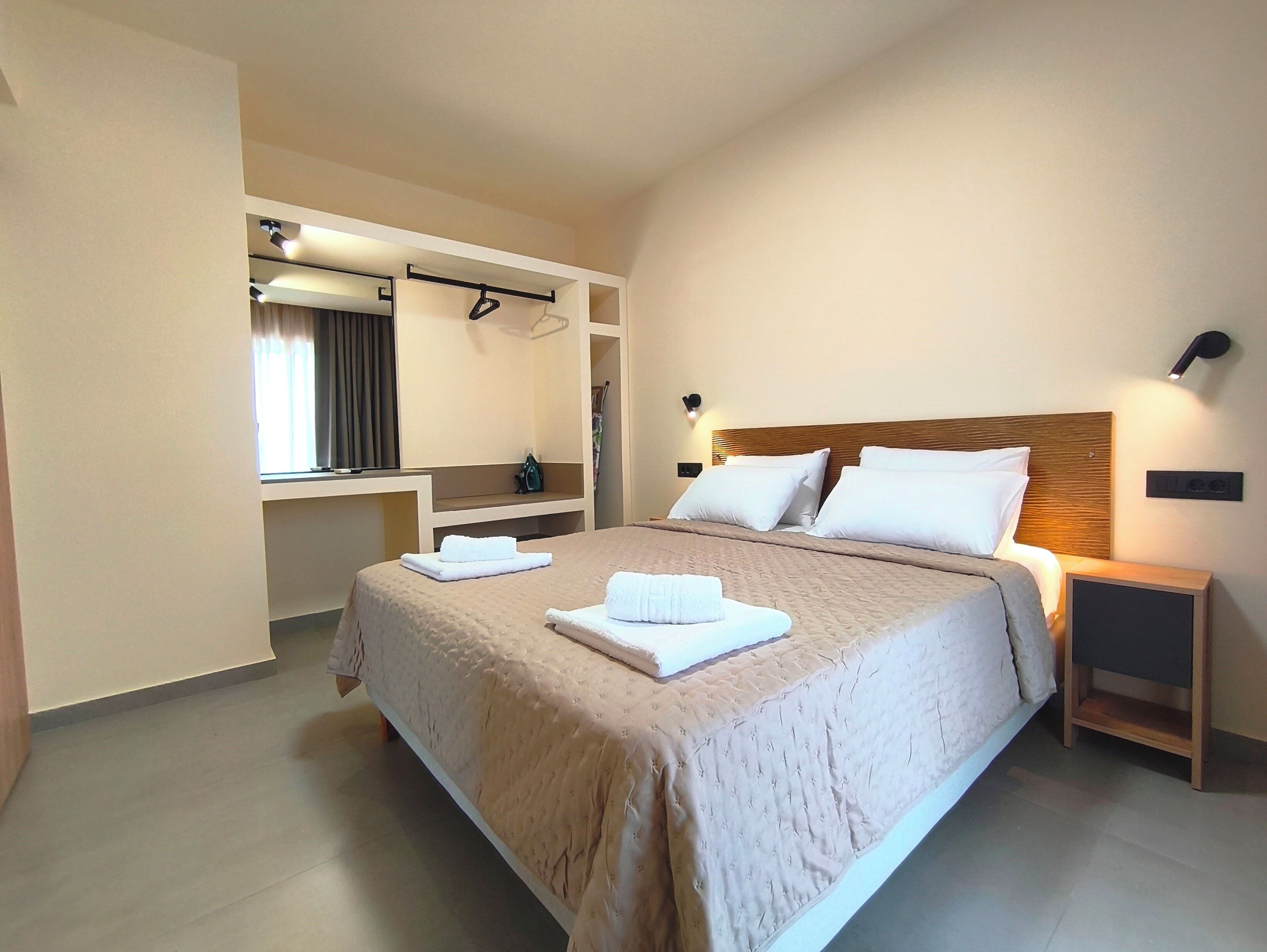 A modern hotel room with a large bed, a built-in wardrobe, a vanity with a mirror, and a bedside table with a lamp.