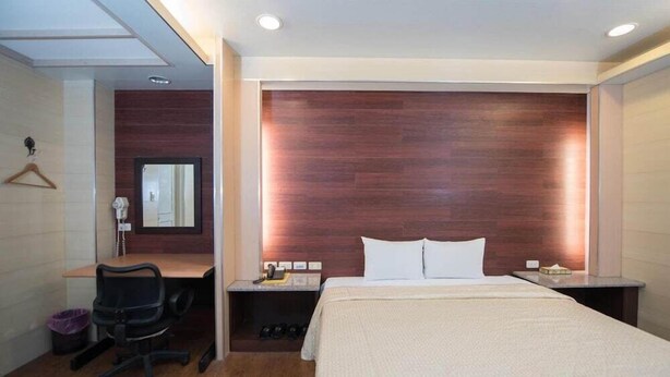 A hotel room with a bed, a desk, a chair, a mirror, and a nightstand. - Empire Hotel (Taipei)