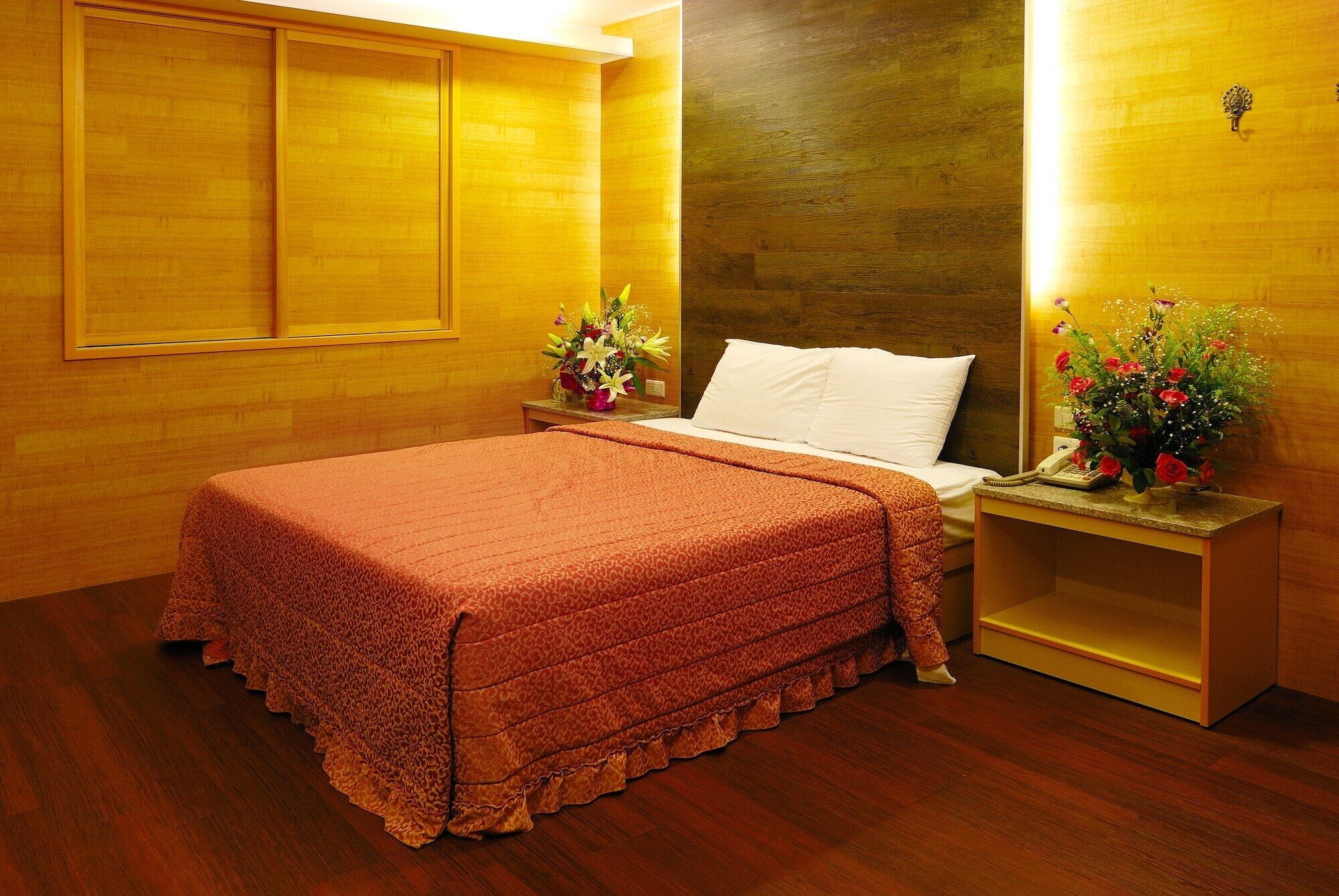 Une chambre d’hôtel avec un plancher en bois, un lit recouvert d’une couvre-lit à motifs, deux tables de chevet ornées de compositions florales et une tête de lit en bois.