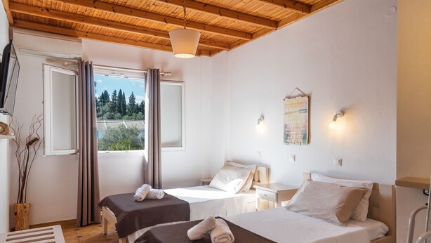 A room with two beds, a wooden ceiling, a window with curtains, a bedside table, and a wall-mounted light fixture. - Three Seasons (Corfu)