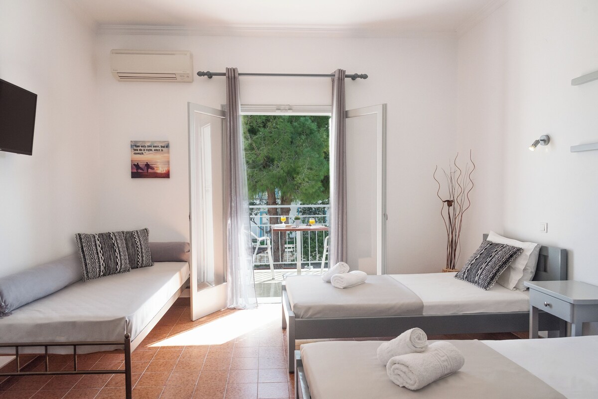 A room with two beds, a sofa, a small table, and a balcony with a view of trees.