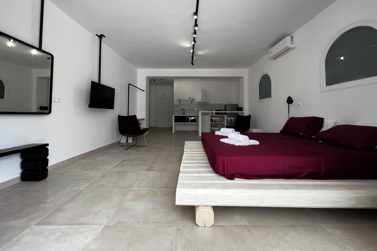 A modern hotel room with a bed, a desk, a chair, a TV, and a kitchen area.