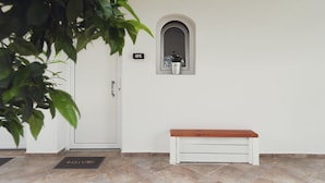 A clean, minimalist entrance with a white door, a welcome mat, a bench, and a small window with a number sign. - Three Seasons (Corfu)