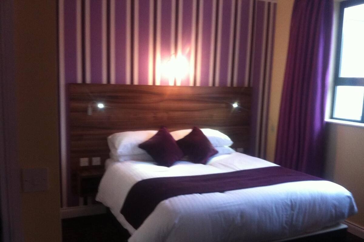 A hotel room with a bed, a headboard, and a window with curtains.