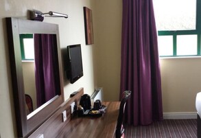 Habitación de hotel con escritorio de madera, televisión de pantalla plana, espejo, ventana con cortinas moradas y una cama.