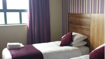 Habitación de hotel con dos camas, cabecera de madera, ventana con cortinas y una toalla sobre la cama.
