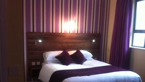Una habitación de hotel con cabecera de madera, cama con ropa de cama blanca y almohadas moradas, y una ventana con cortinas moradas.