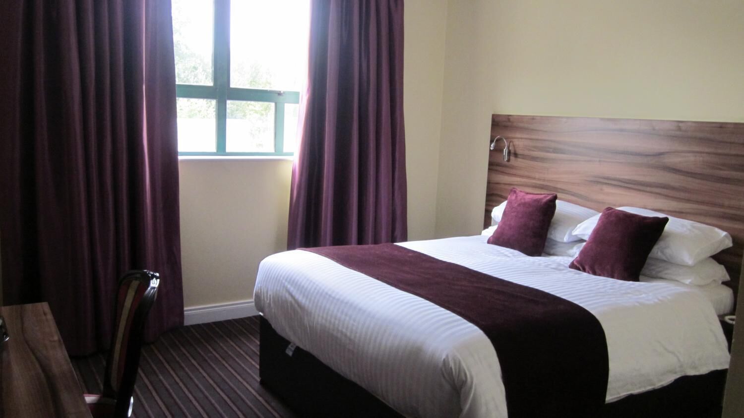 A hotel room with a large bed, dark curtains, a wooden headboard, and a window with a view.
