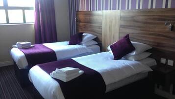 Habitación de hotel con dos camas, cada una con una manta y almohadas moradas, y toallas sobre las camas.