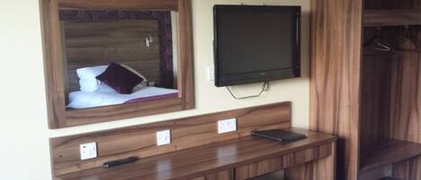 A room with a wooden desk, a mirror, a flat-screen TV, and a wardrobe.