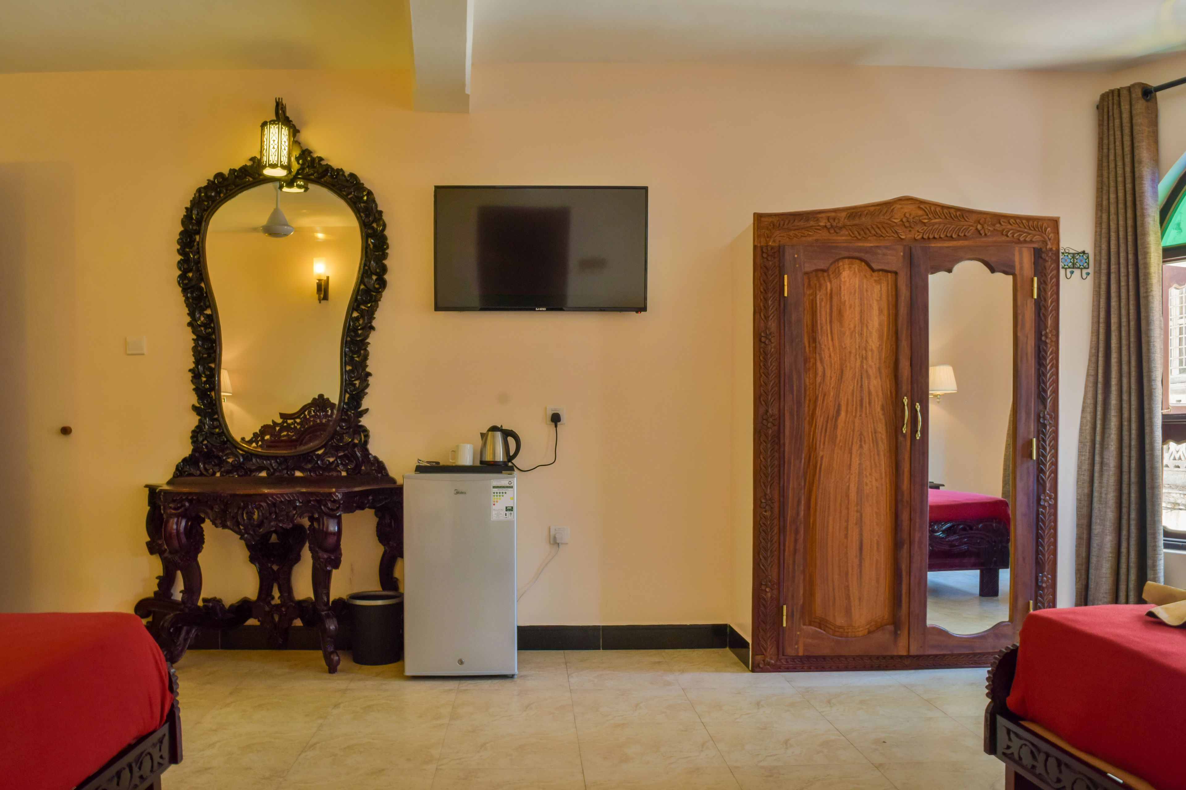 A hotel room with a large mirror, a television, a wardrobe, a small refrigerator, and two beds with red bedding.