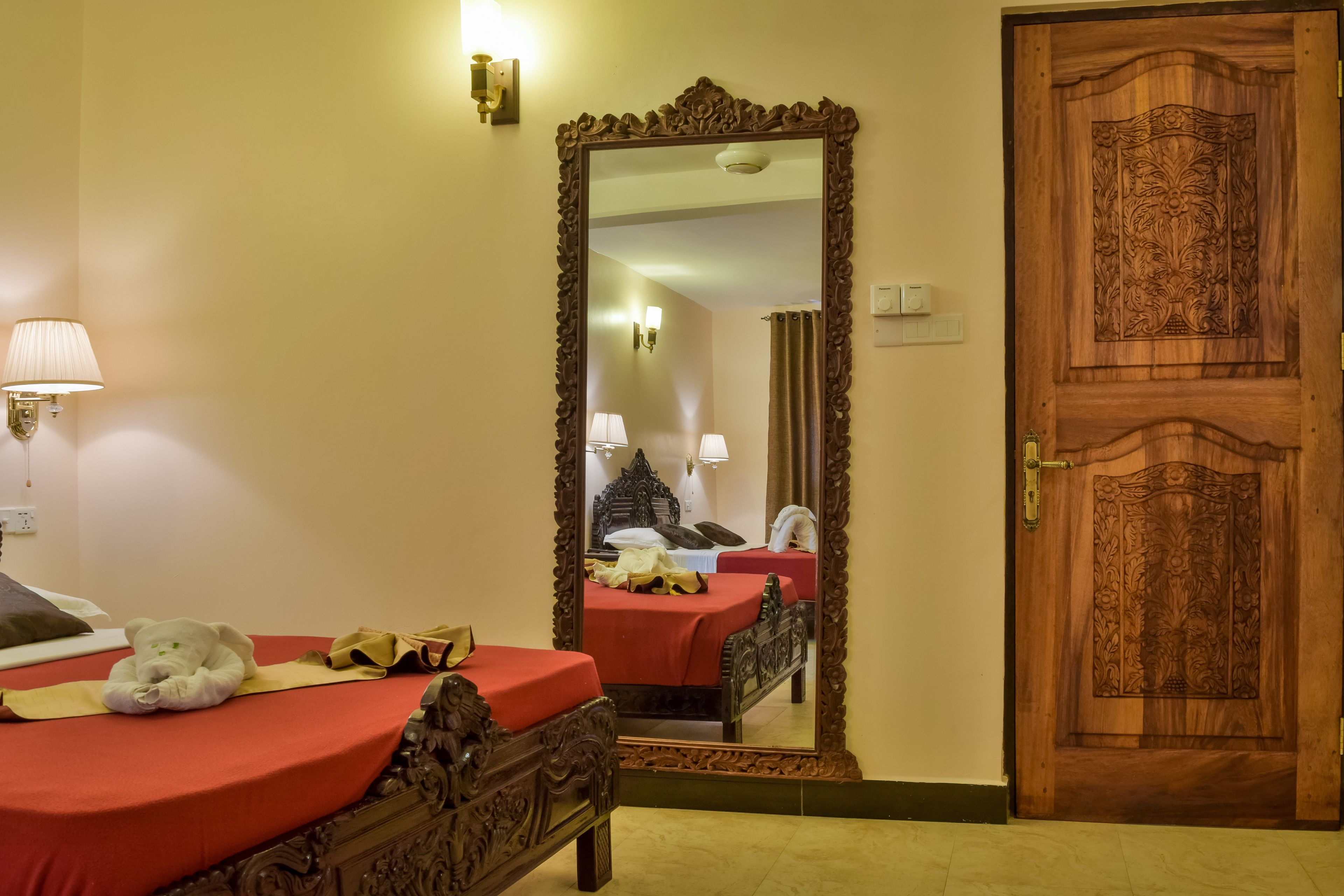 A hotel room with a wooden bed, a large mirror, a wooden door, and a red bedspread.