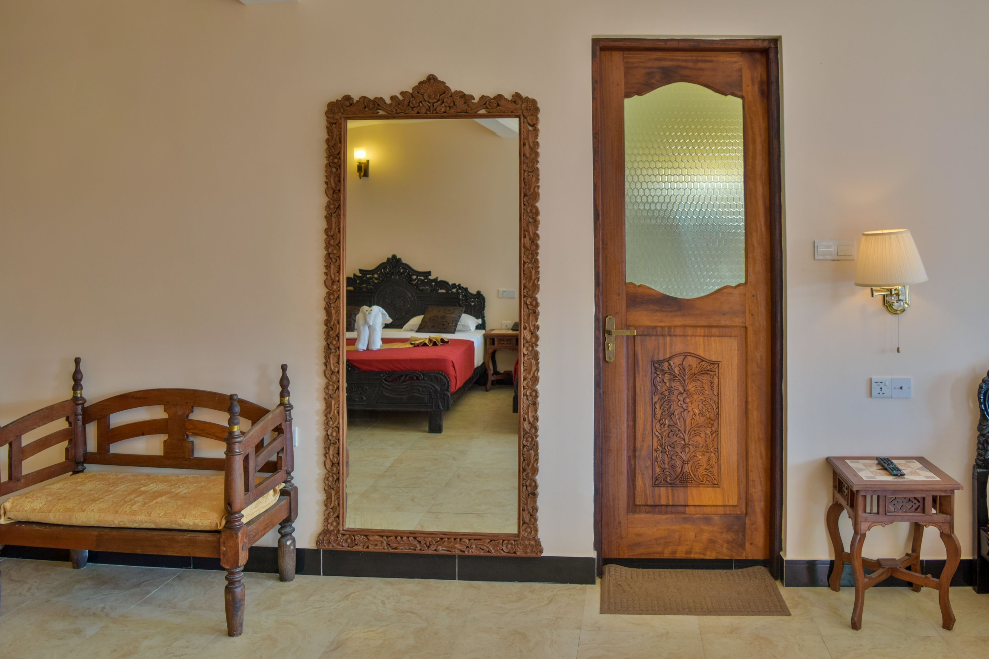 A room with a wooden bed, a wooden chair, a mirror, a door with a frosted glass panel, and a small table with a lamp.