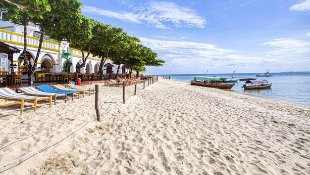 A beach with sun loungers and a building with arches. - Tembo Palace Hotel (Zanzibar Town)