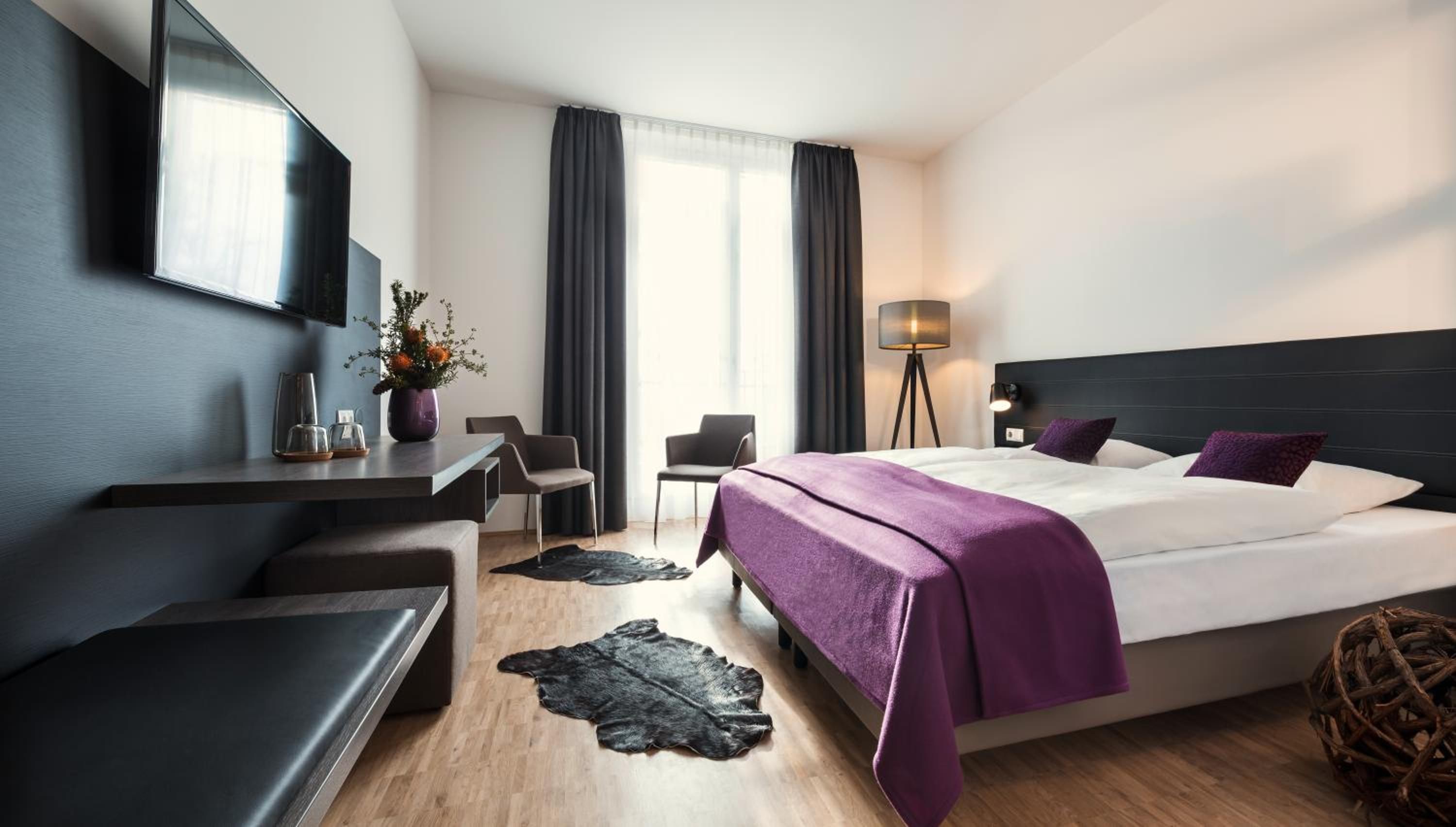 A modern hotel room with a bed, a desk, a chair, a lamp, and a large window.