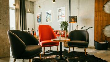 A modern interior with two dark armchairs, a red chair, and a round wooden table. There are abstract paintings on the wall and a potted plant.