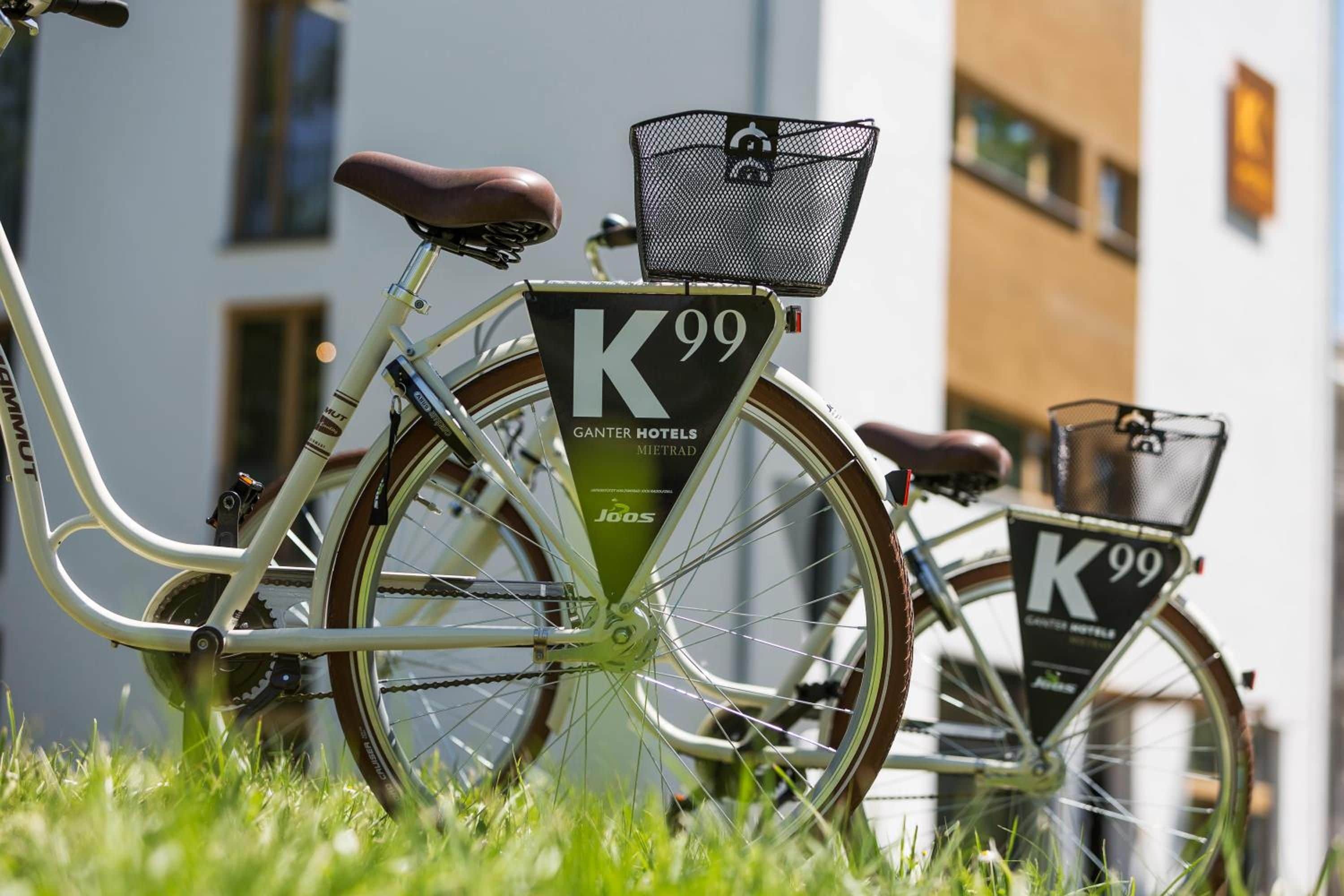 A bicycle with a 'K99' sign and a 'Ganter Hotels' label, parked on grass with buildings in the background.