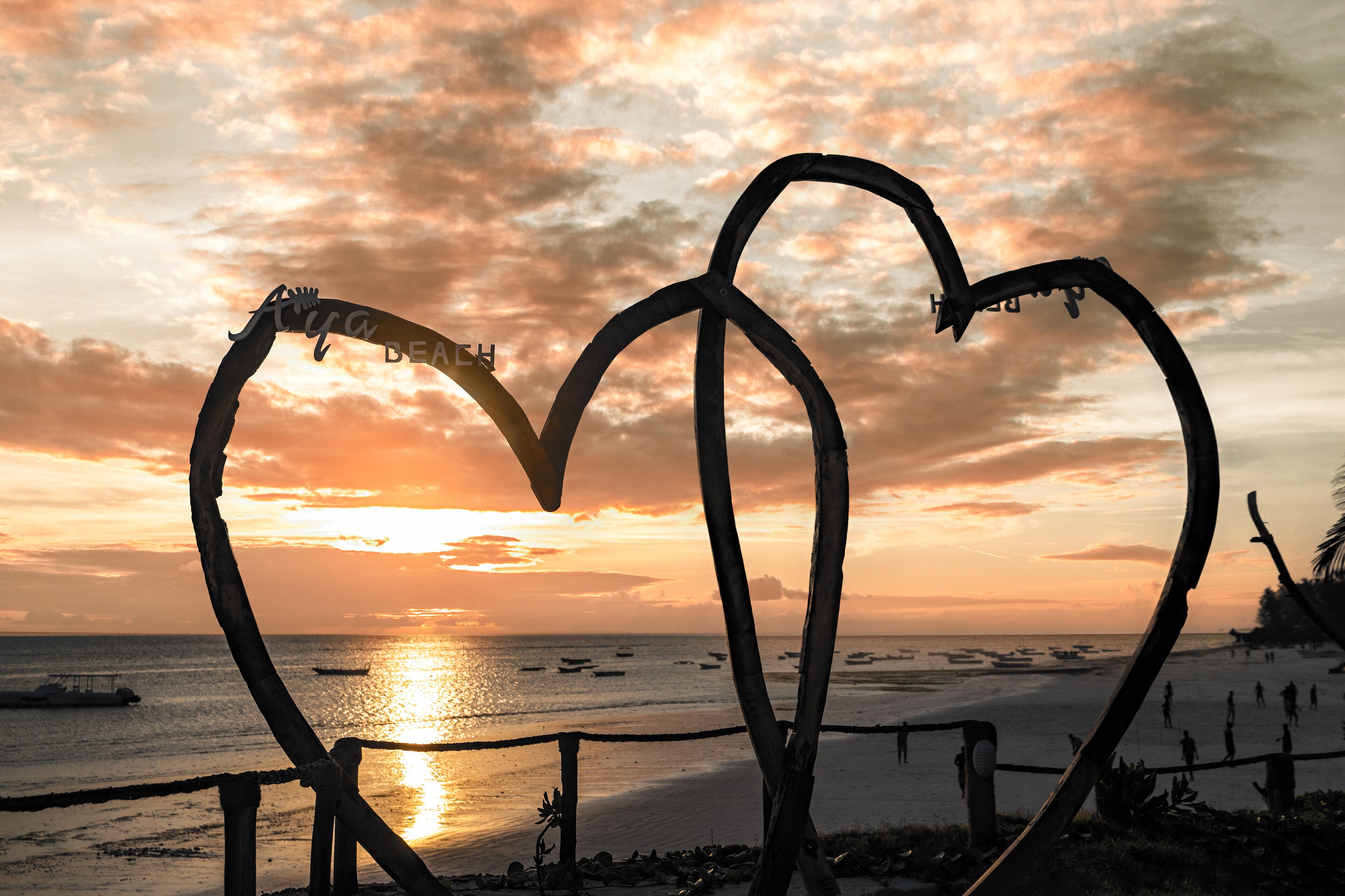 Escultura de metal em formato de coração numa praia, com um pôr do sol ao fundo.