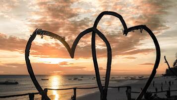 Escultura de metal em formato de coração numa praia, com um pôr do sol ao fundo.