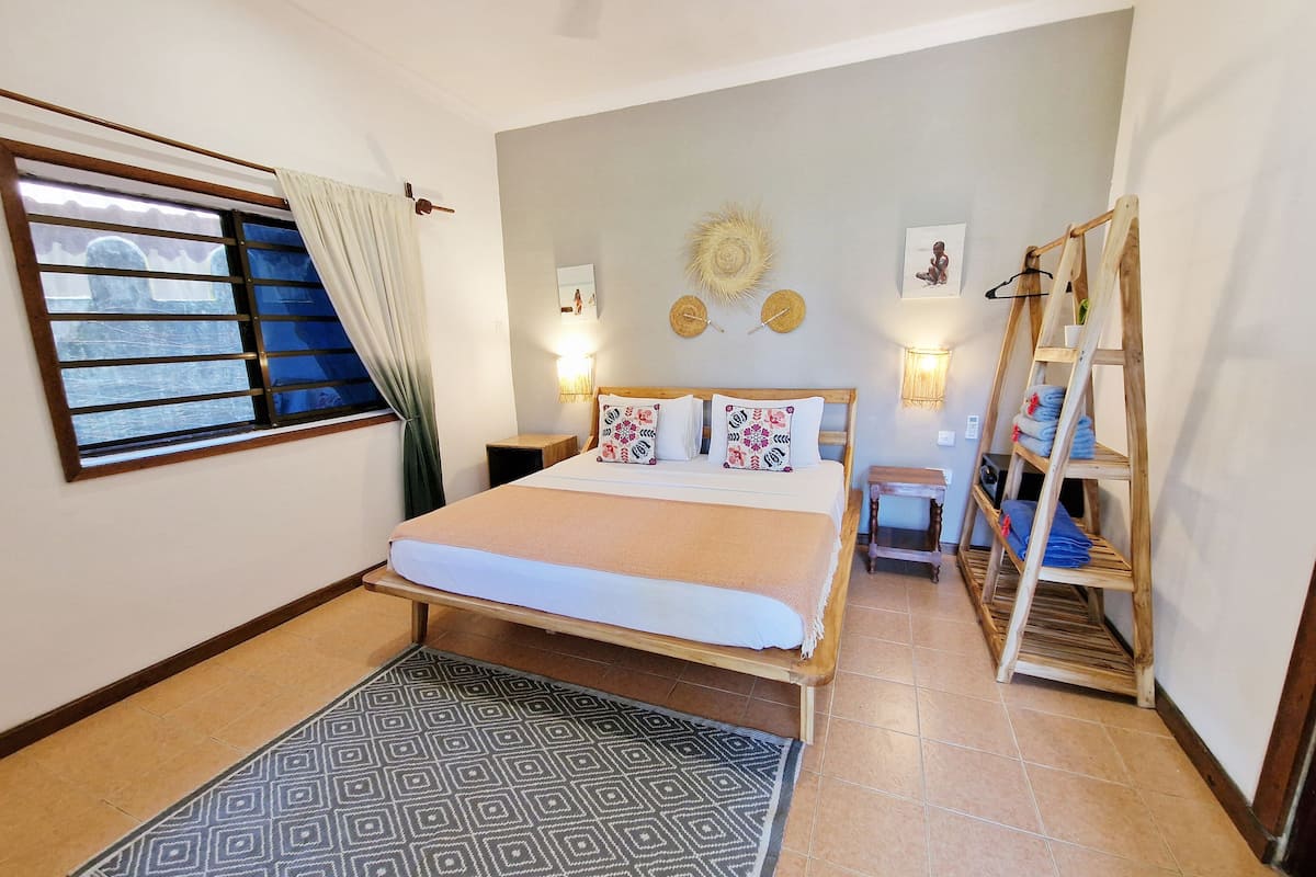 A bedroom with a bed, a window with curtains, a bedside table, a wall-mounted lamp, a small shelf, and a wooden ladder.