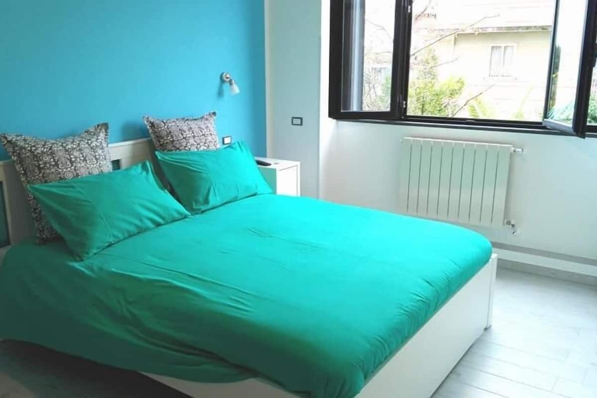 Une chambre à coucher avec un lit, deux oreillers, une table de chevet, une fenêtre et un radiateur.