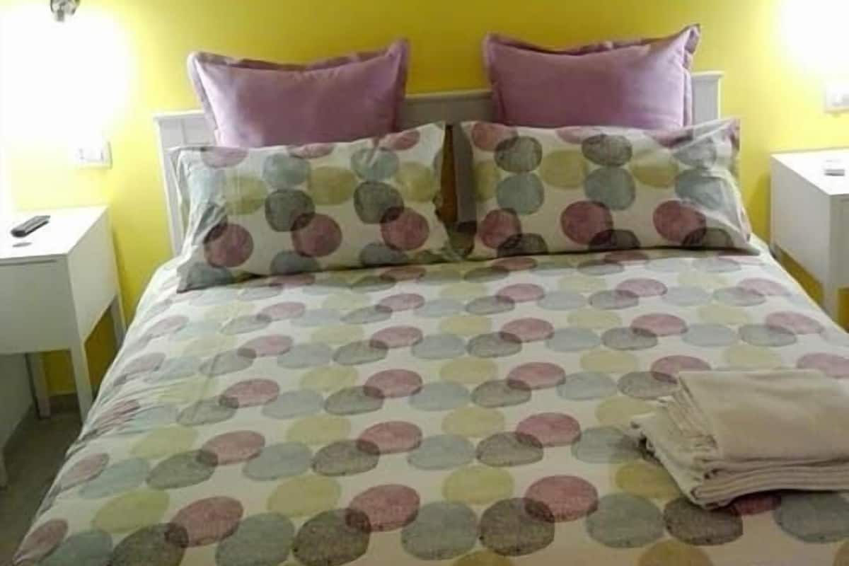 Un lit avec une couvre-lit à pois et deux oreillers, une table de chevet blanche avec une lampe, et un mur jaune.