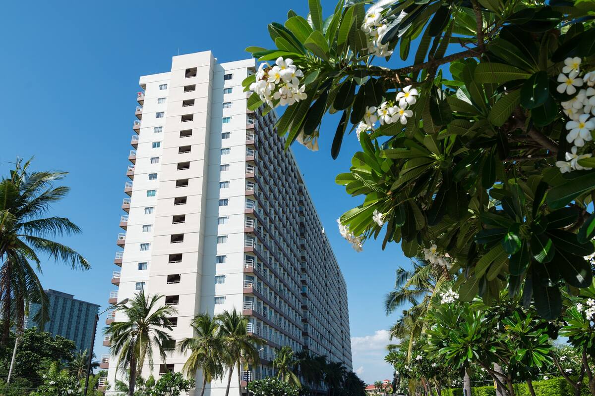 A tall, modern hotel with multiple floors and balconies, surrounded by lush greenery and palm trees.