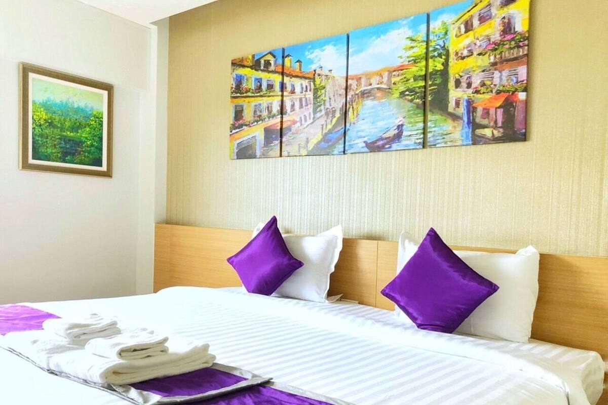 A hotel room with a large bed, purple pillows, and a painting of a canal scene on the wall.