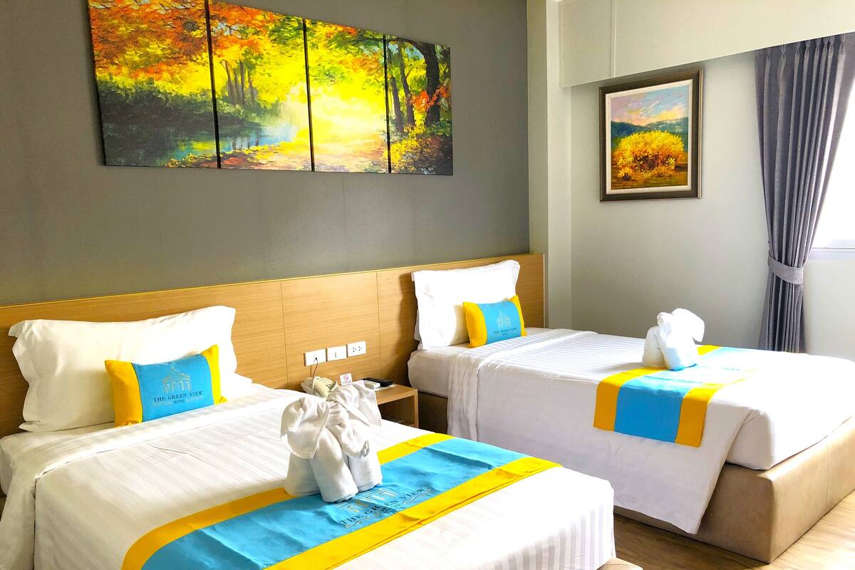 A hotel room with two beds, each with white bedding and blue and yellow accents. There are two paintings on the wall, one depicting a forest scene and the other a landscape.