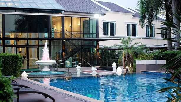 A swimming pool with a fountain, surrounded by lounge chairs and a building with large windows and a staircase. - The Green View Hotel (Bang Sao Thong)