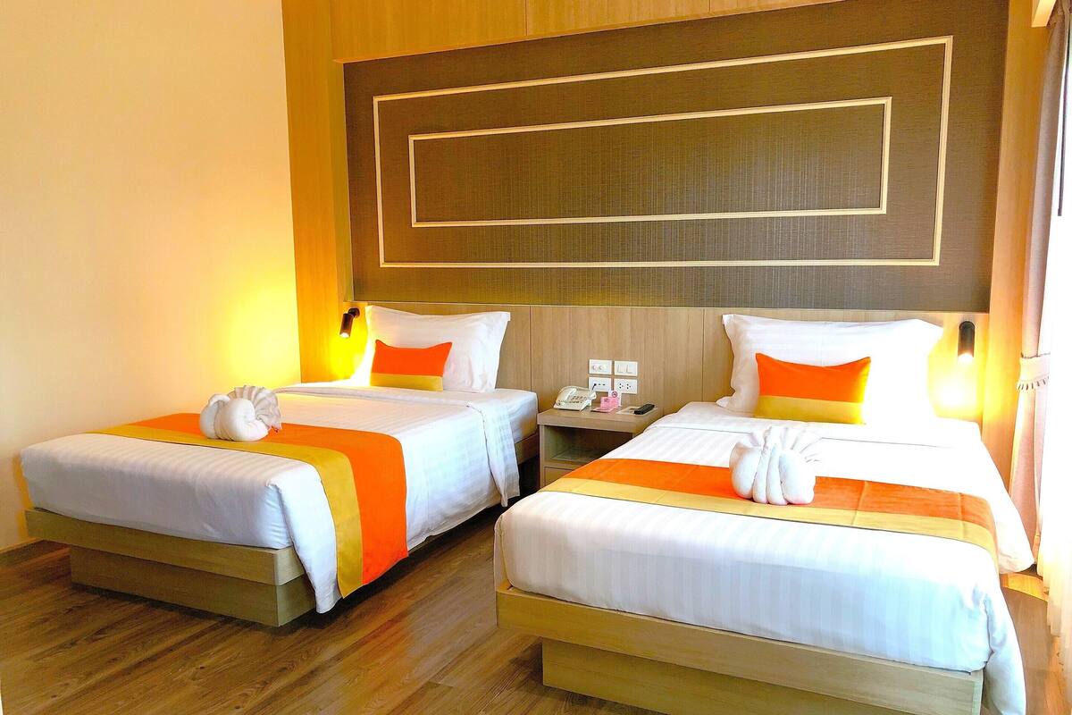 Two double beds with white and orange bedding, wooden headboards, bedside tables with lamps, and a wooden floor.