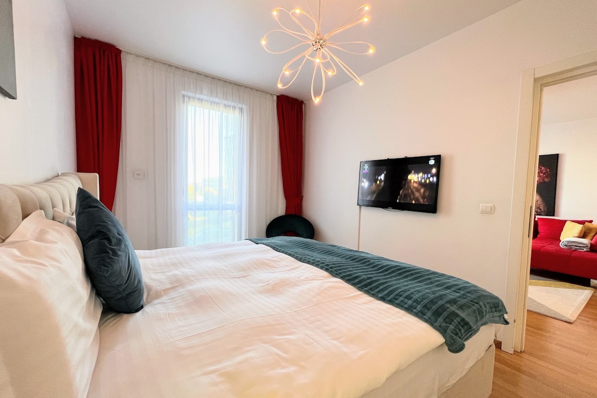 Une chambre avec un lit, un téléviseur fixé au mur et un canapé rouge dans la pièce adjacente.