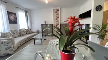 Una sala de estar moderna con un sofá, una mesa de centro de vidrio, una planta en maceta roja, un televisor y un reloj de pared.