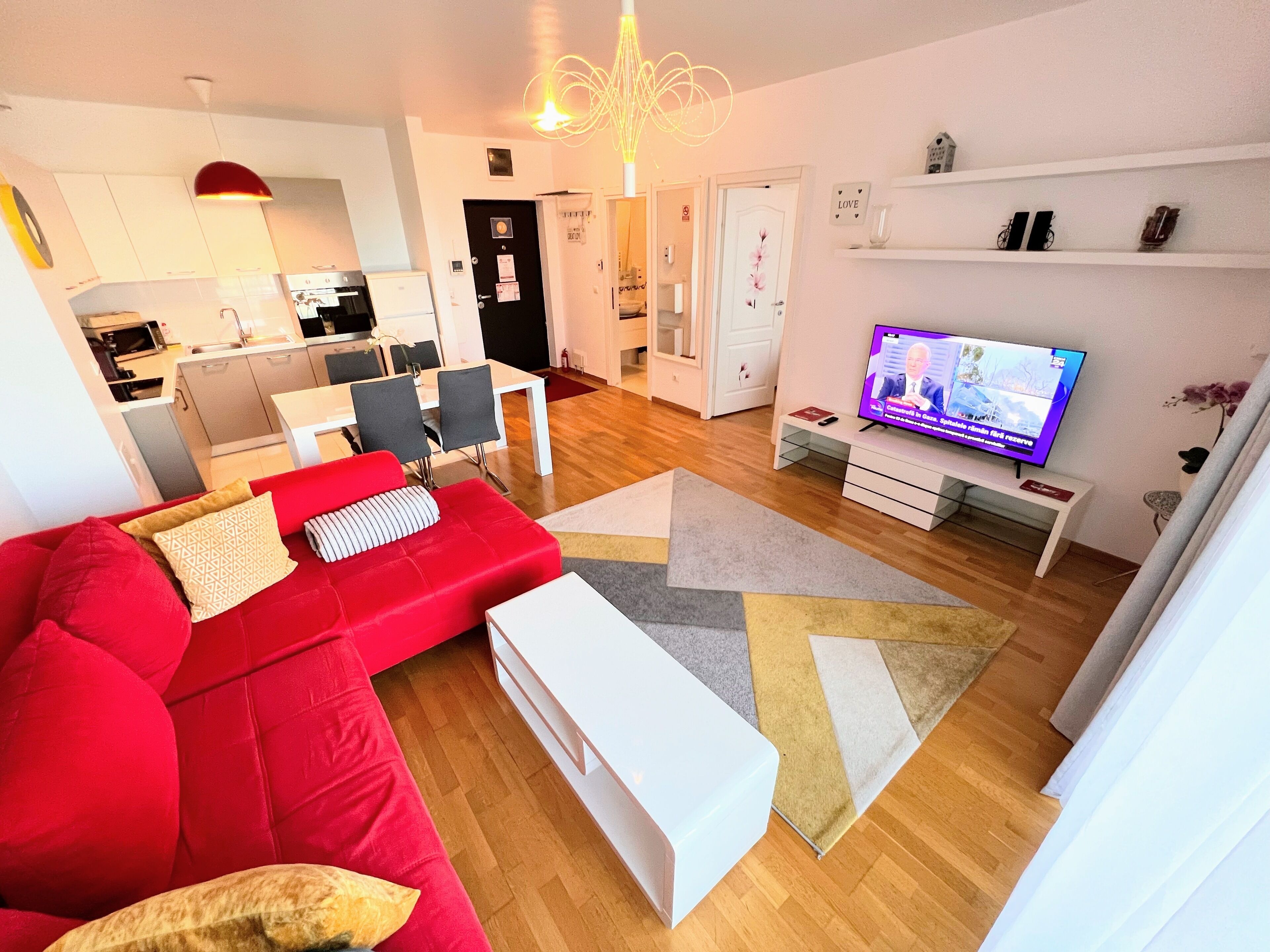 A modern living room with a red sofa, a white coffee table, and a TV on a stand. There is a kitchen in the background with white cabinets and a dining area with a table and chairs.