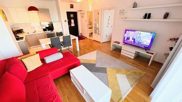 A modern living room with a red sofa, a white coffee table, and a TV on a stand. There is a kitchen in the background with white cabinets and a dining area with a table and chairs.