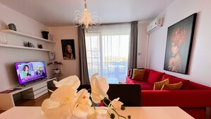 Uma sala de estar moderna com um sofá vermelho, uma mesa branca com um vaso de orquídeas, uma TV exibindo um canal de notícias e uma janela grande com cortinas.