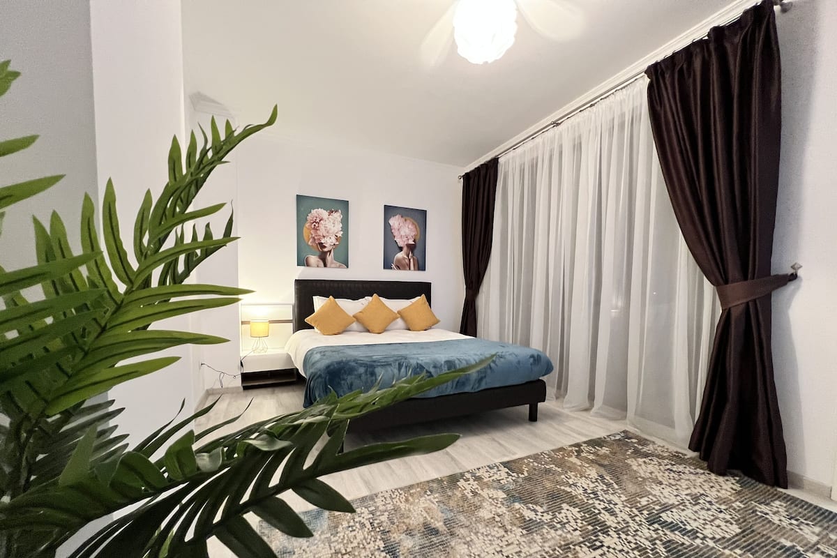 Une chambre à coucher moderne avec un lit, une table de chevet, une lampe et une grande fenêtre avec des rideaux.