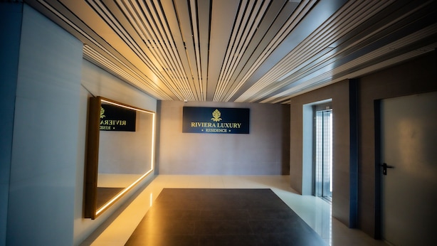 Um corredor moderno com piso escuro, teto com luminárias lineares e uma placa indicando "RIVIERA LUXURY RESIDENCE".