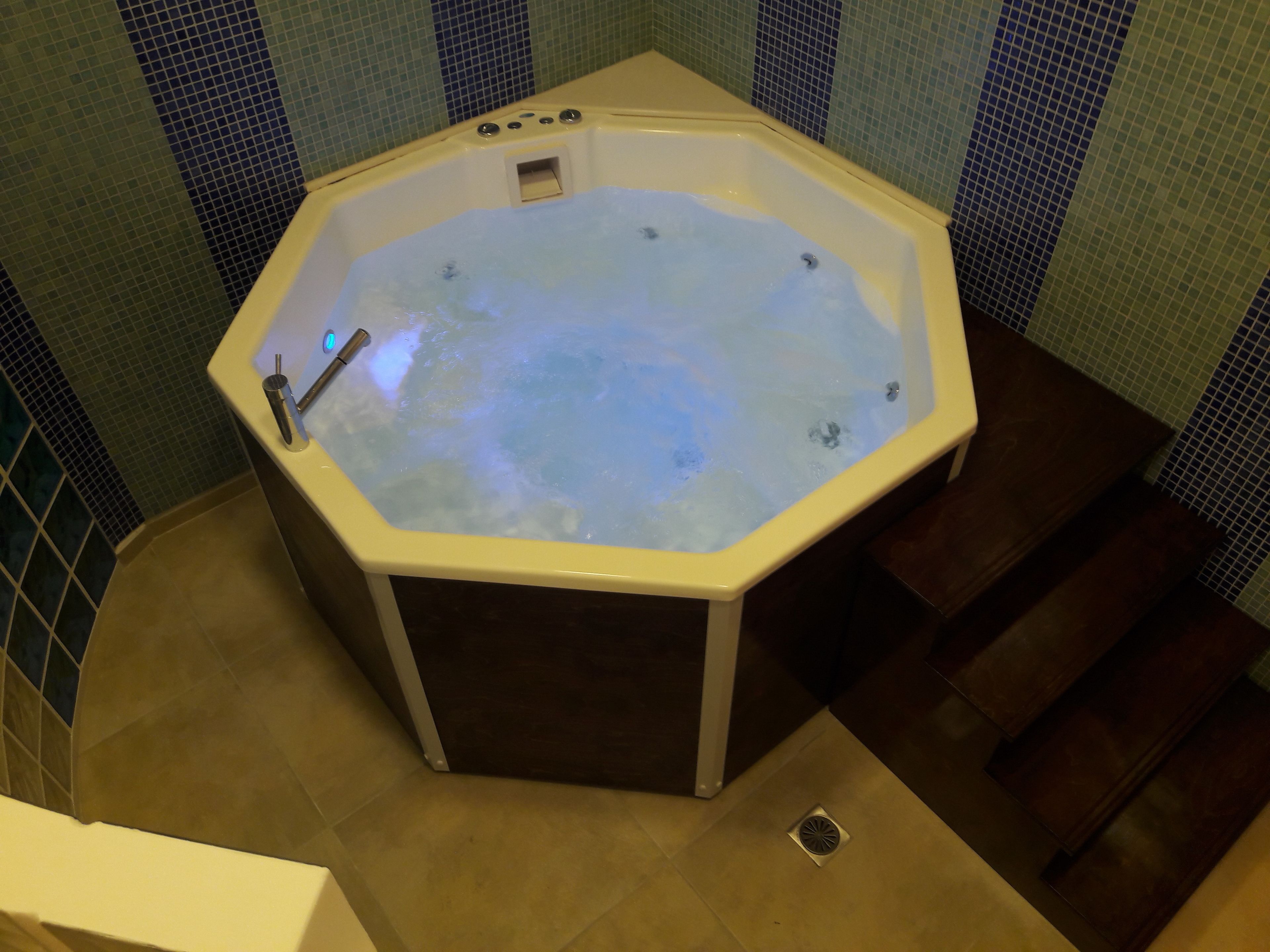A hot tub in a corner with a tiled wall and a wooden floor.